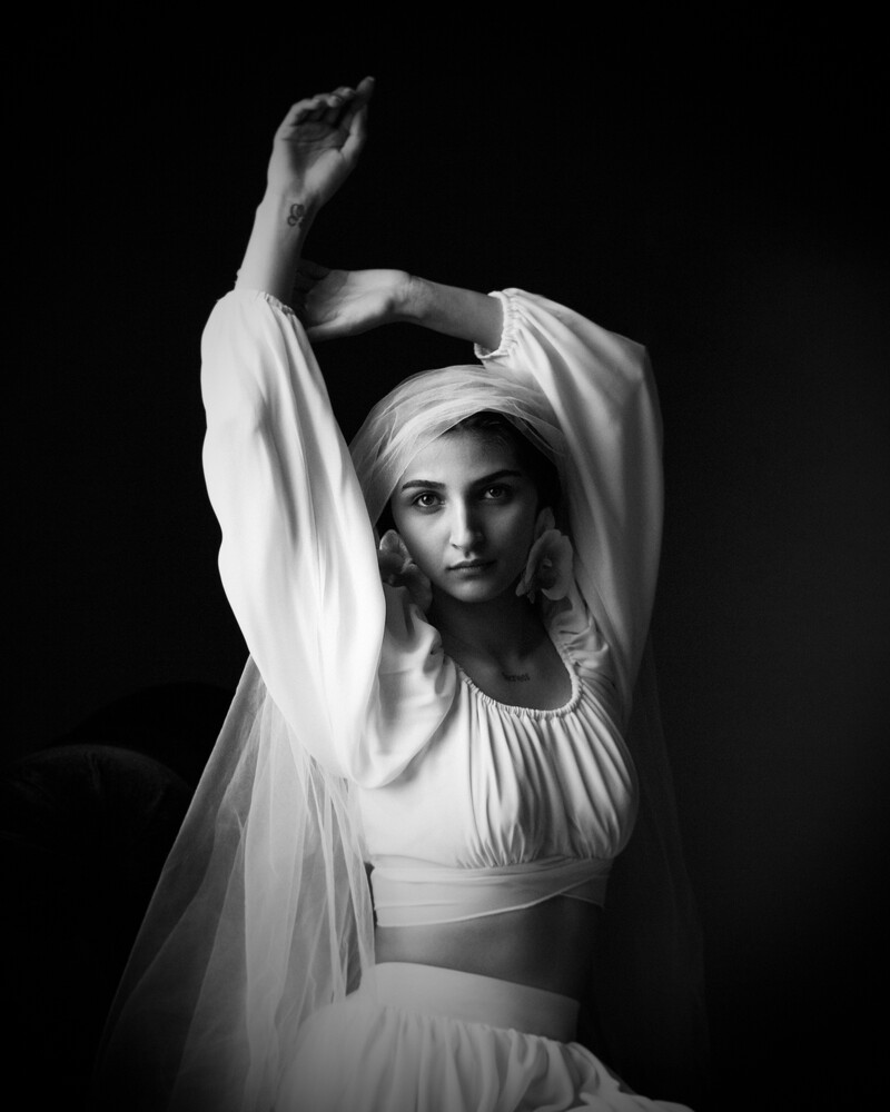 Black and white portrait of a woman with raised arms, wearing a white head wrap and flowing sheer sleeves against a black background.