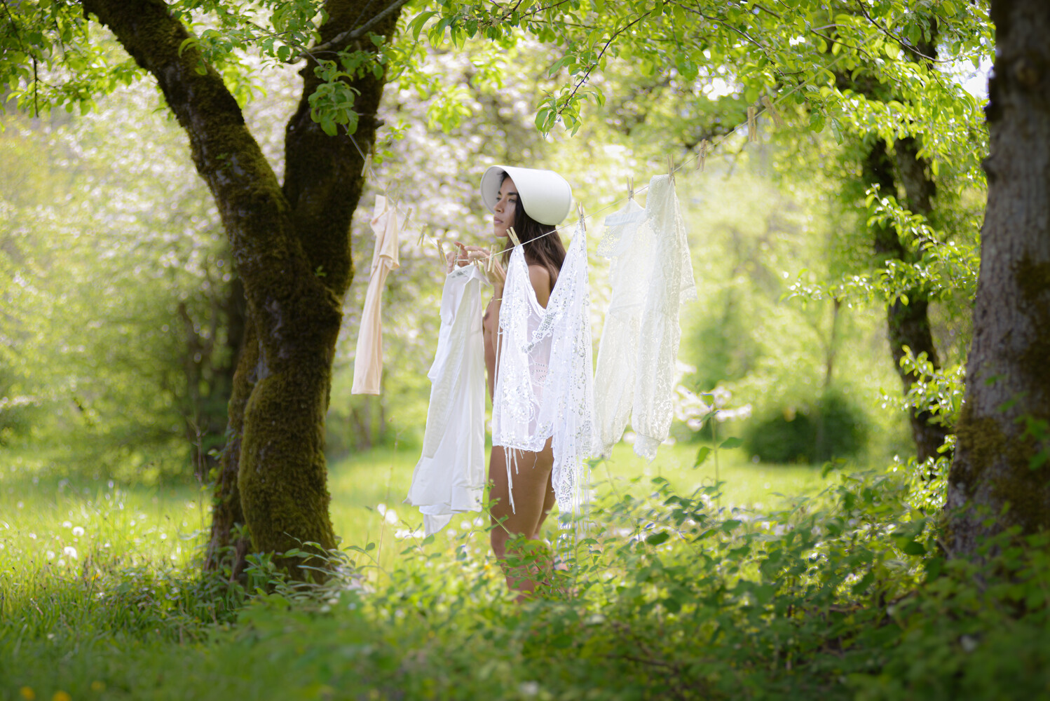 Woman in a white bonnet holding flowing white fabric among mossy trees in a sunlit green orchard