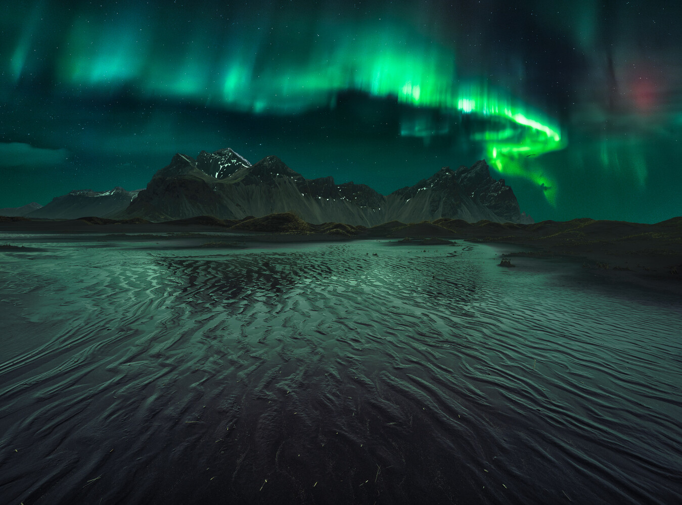 Northern lights dancing in green waves above snow-covered mountains and black sand beach