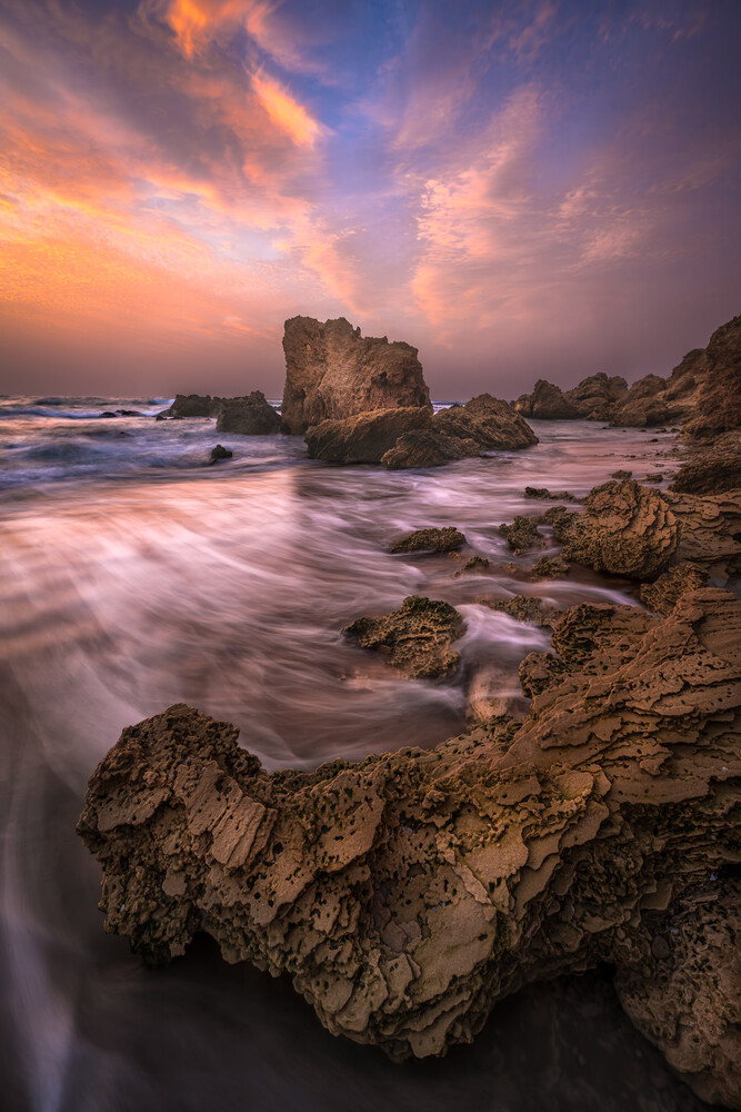 Rocky coastal landscape at sunset with smooth water and dramatic purple-orange sky