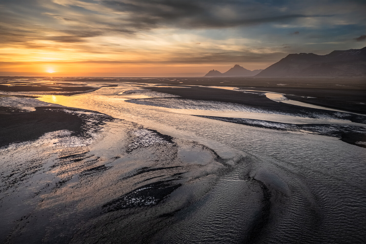 Aerial view of braided river channels winding across a dark floodplain at sunset, with mountains on the right.