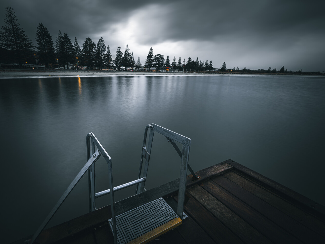 Metal dock with railings extending into a calm lake under stormy skies and distant evergreen treeline