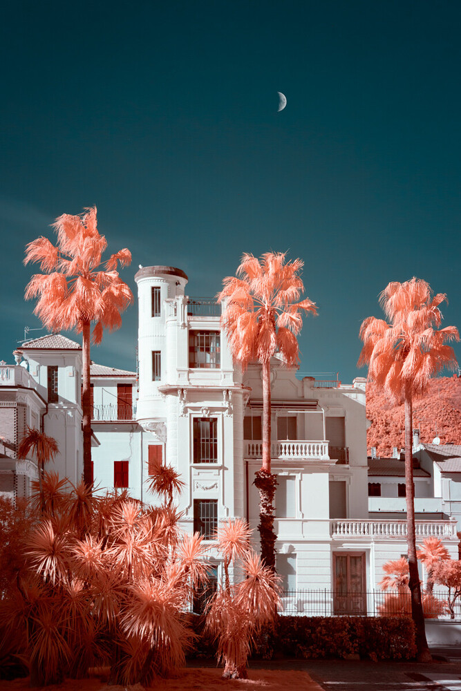 White Mediterranean-style building with palm trees in infrared against teal sky with crescent moon