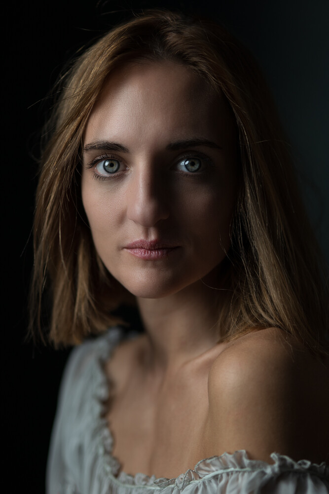 Close-up portrait of a woman with shoulder-length light brown hair and blue eyes, gazing toward the camera against a dark background.