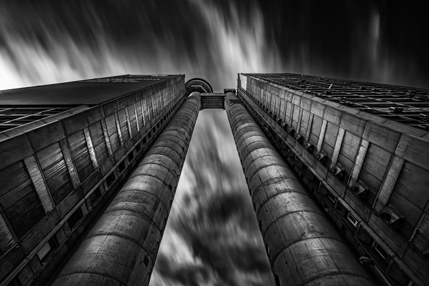 Low-angle black and white view looking up between two tall concrete towers joined by a walkway, with streaked clouds overhead.