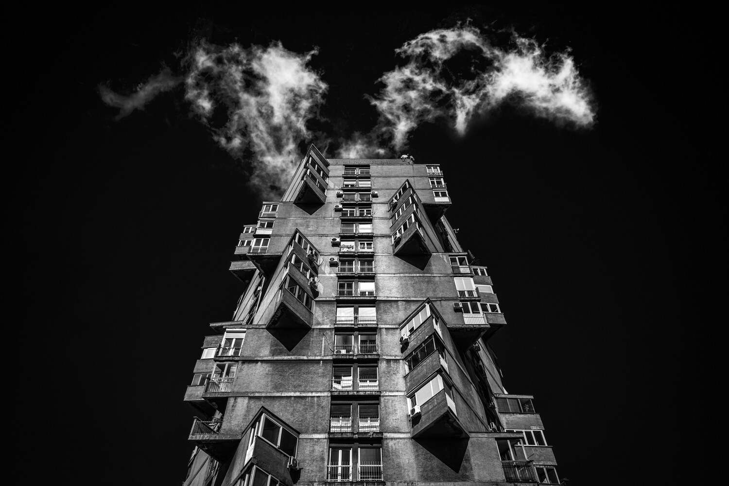 Low-angle black and white view of a concrete apartment tower with staggered angular balconies under a dark, cloudy sky