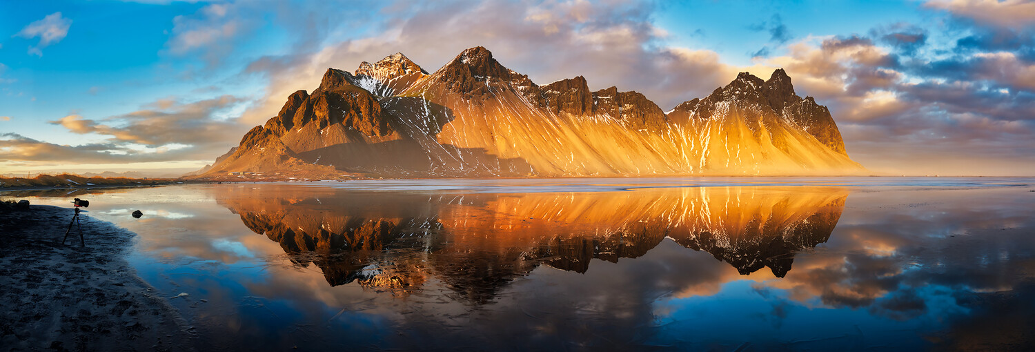 Golden-lit mountain peak reflected in still water at sunrise with photographer and tripod on shore