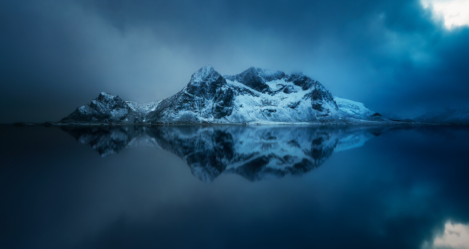Snow-covered mountain peaks reflected in still dark water under stormy blue sky