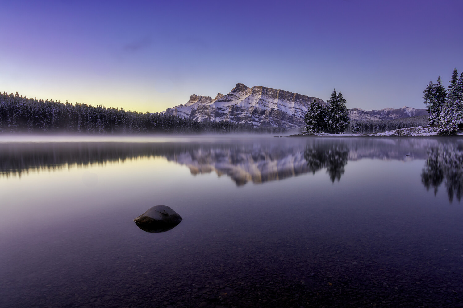 Snow-capped mountain reflected in still lake at dawn with forested shoreline