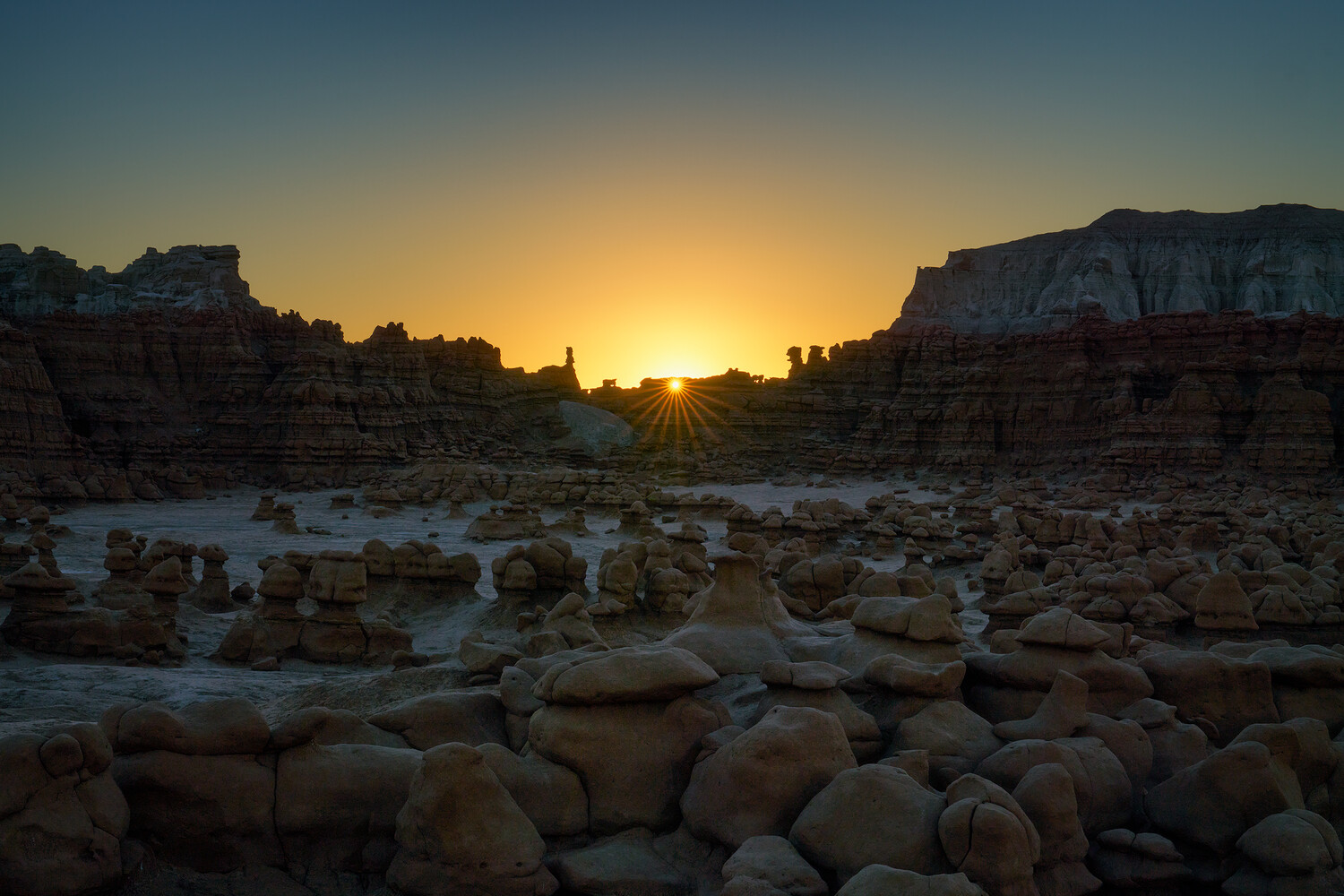 Sun setting behind eroded sandstone hoodoos and cliffs in a desert landscape, with a starburst of rays over the ridge.