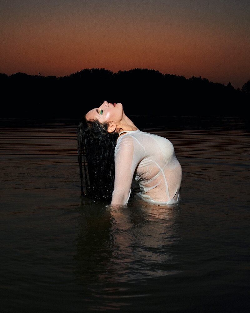 Woman in light-colored dress kneeling in water at dusk with trees silhouetted against dark sky