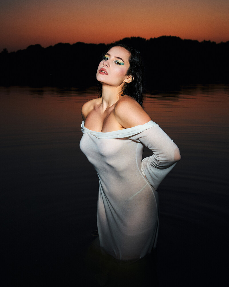 Woman in white off-shoulder dress posing in calm water at sunset