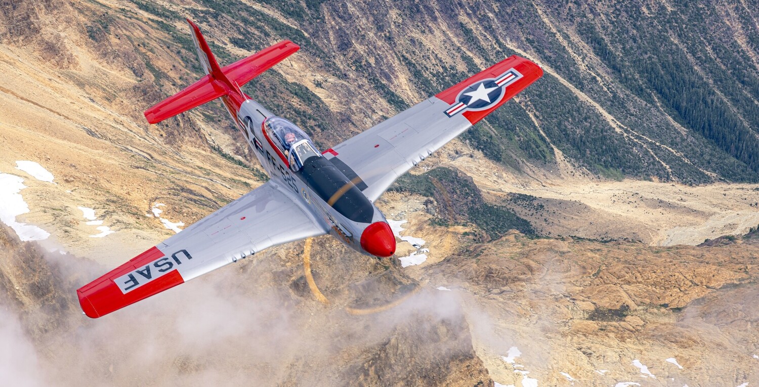 Red and white aerobatic aircraft in steep bank over mountainous terrain with snow patches