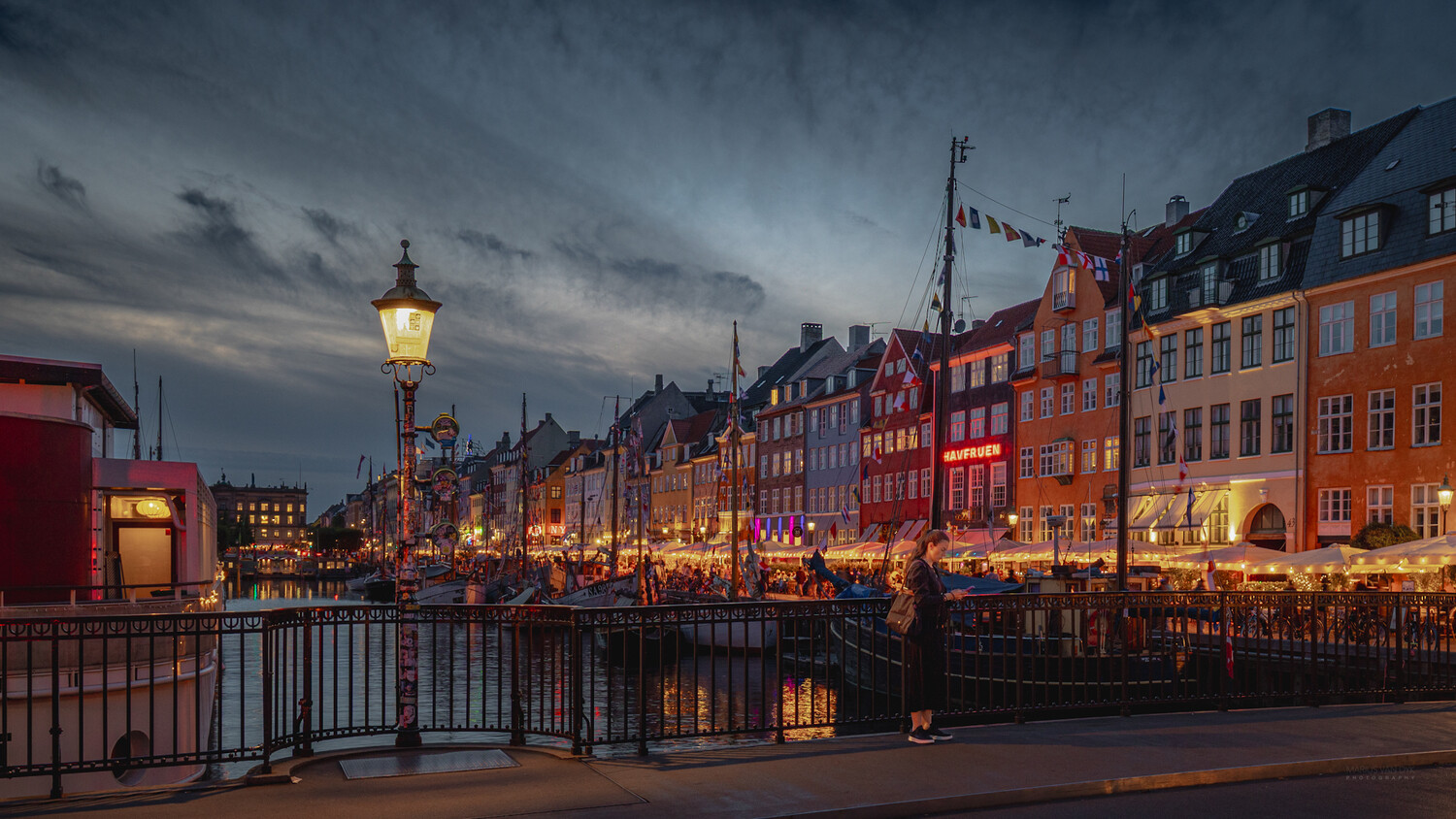 Colorful waterfront buildings illuminated at dusk along a European canal with bridge and street lamp