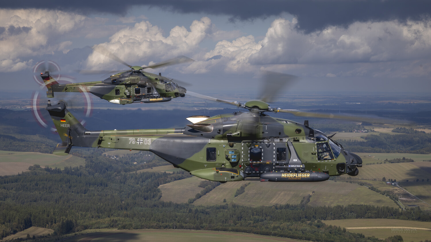 Two military transport helicopters in formation flying over forested and agricultural landscape