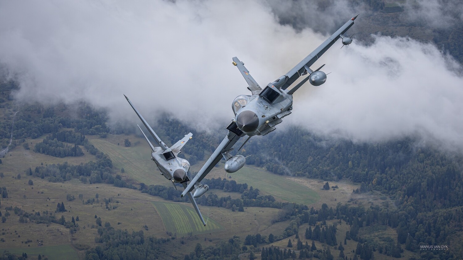 Two military transport aircraft flying in formation above forested mountainous terrain and clouds
