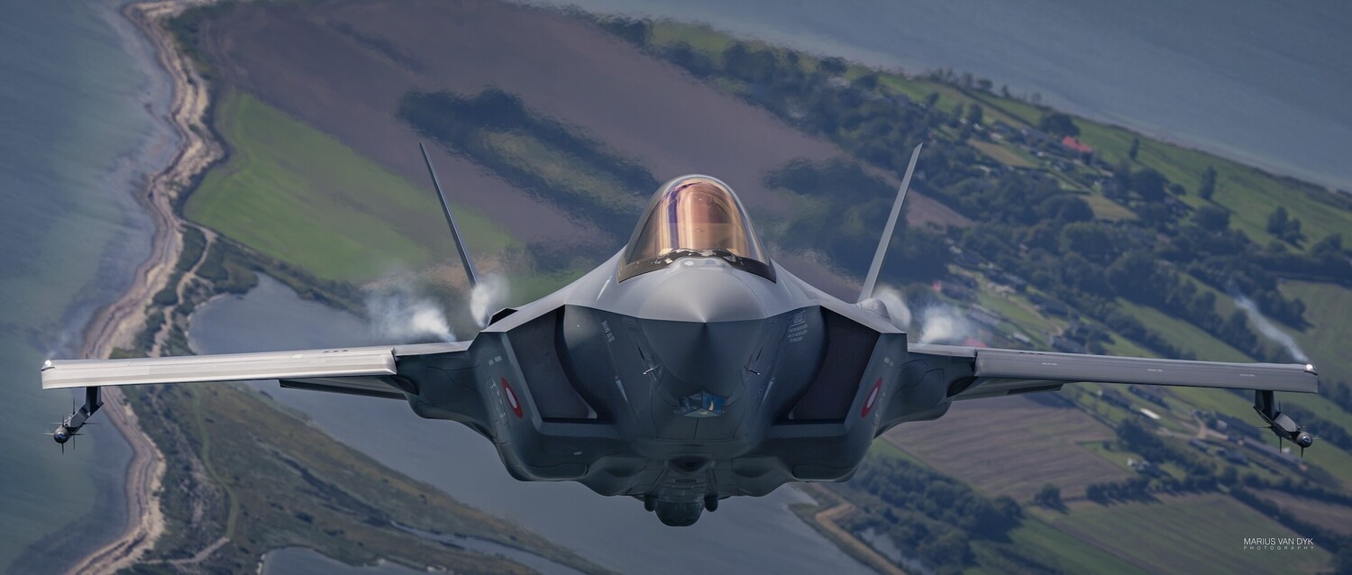 F-35 Lightning II fighter jet in head-on flight over patchwork farmland and coastline