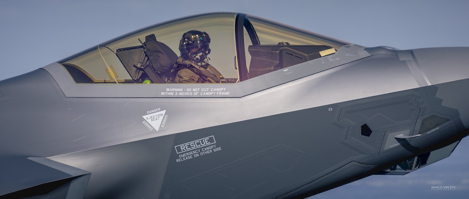 Fighter jet cockpit with pilot wearing flight helmet visible through canopy against blue sky