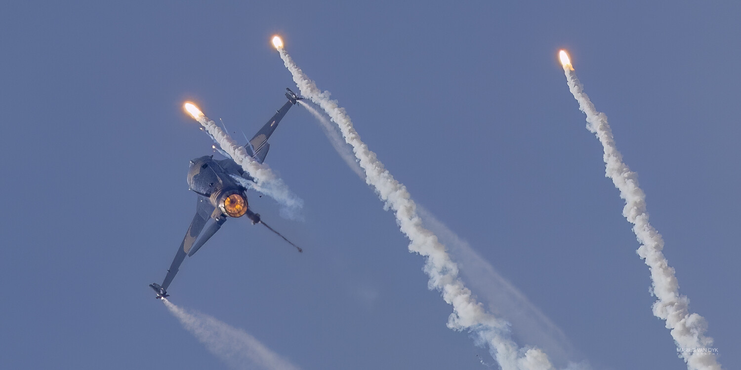 Military fighter jet performing aerial maneuver with afterburner flares deployed against clear blue sky