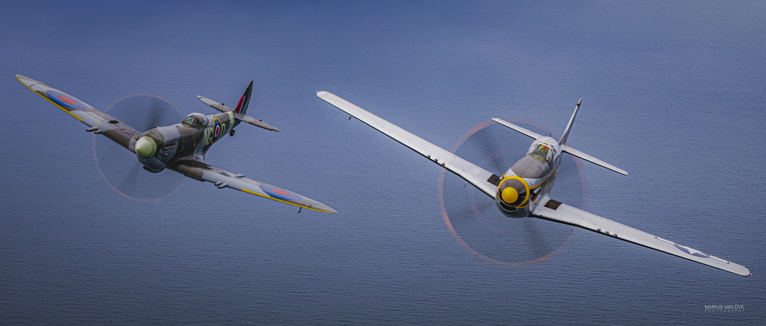 Two vintage WWII fighter planes, a camouflaged Spitfire and a silver fighter with a yellow spinner, flying in formation over calm blue sea