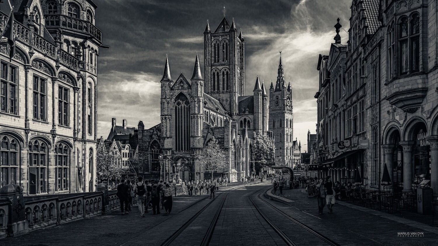 Black and white view of a Gothic church with spires and a tall belfry at the end of a cobbled street with tram tracks.