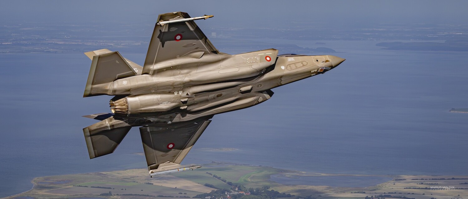 Gray F-35 Lightning II fighter jet banking over blue sea and green coastline, photographed from another aircraft
