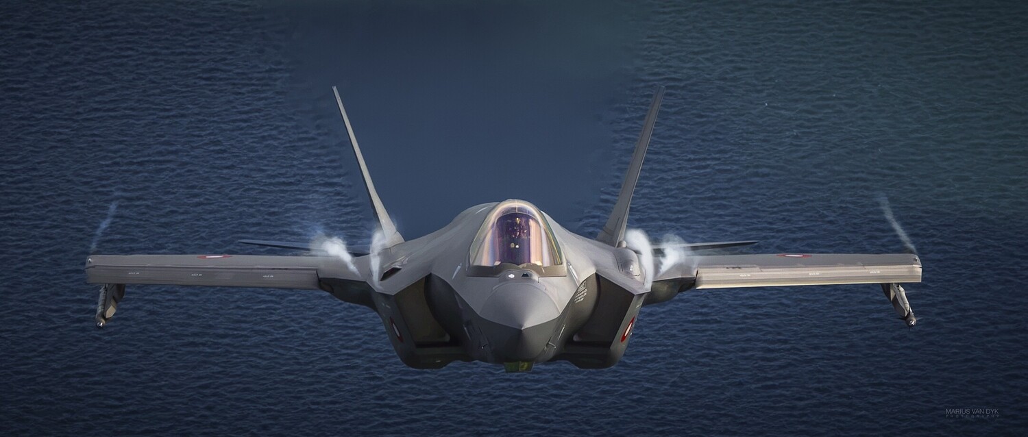 Head-on view of a gray F-35 Lightning II fighter jet flying low over dark blue ocean, with vapor curling off its wings.