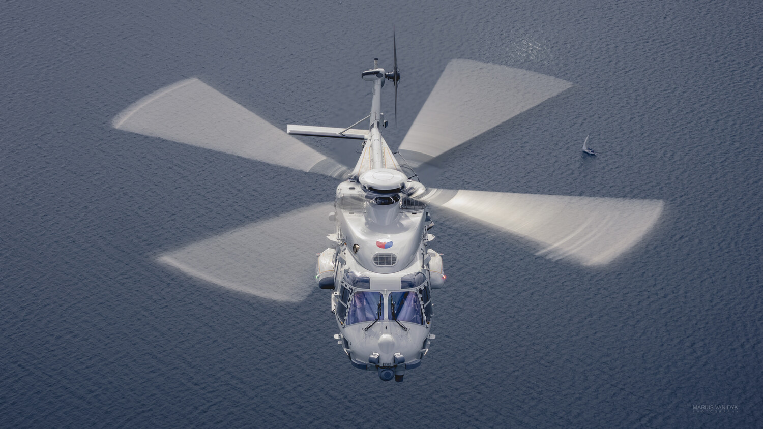 Aerial view of a white helicopter with blurred rotor blades hovering above calm, dark blue water, with a small boat in the distance.