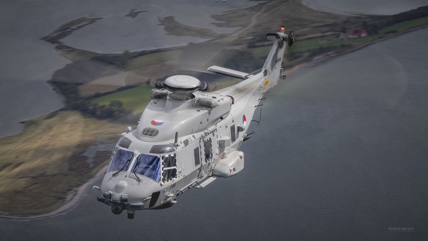 Grey naval helicopter in flight over a coastline of green fields and water, photographed from the air at a three-quarter angle.