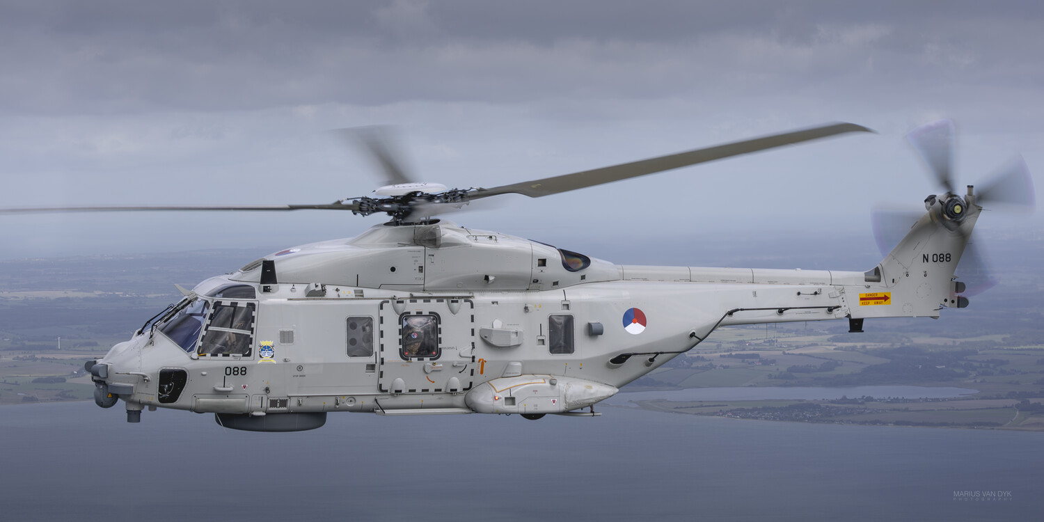 Grey military helicopter in flight over water and coastal farmland, showing a Dutch roundel and side number 088