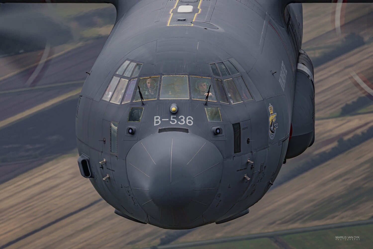 Grey military transport aircraft flying head-on over farmland, nose marked B-536, with crew visible through the cockpit windows