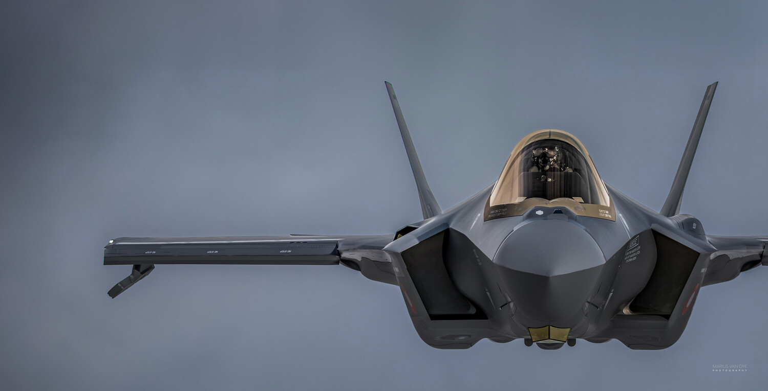 Gray military fighter jet in flight, seen nose-on from slightly below, with its canopy and twin vertical tails visible against an overcast sky.
