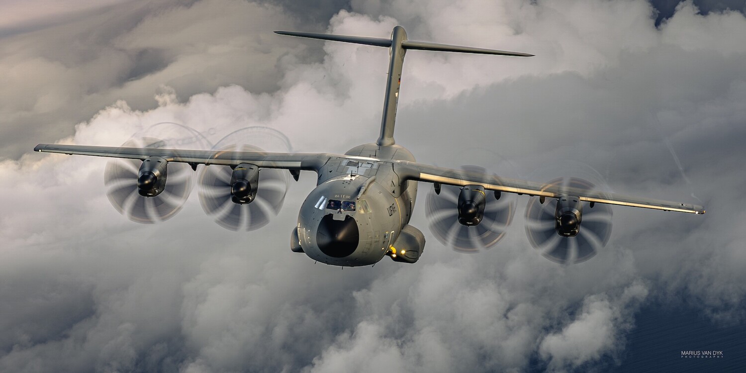 Gray military turboprop transport plane with four spinning propellers flying above clouds, seen from the front.