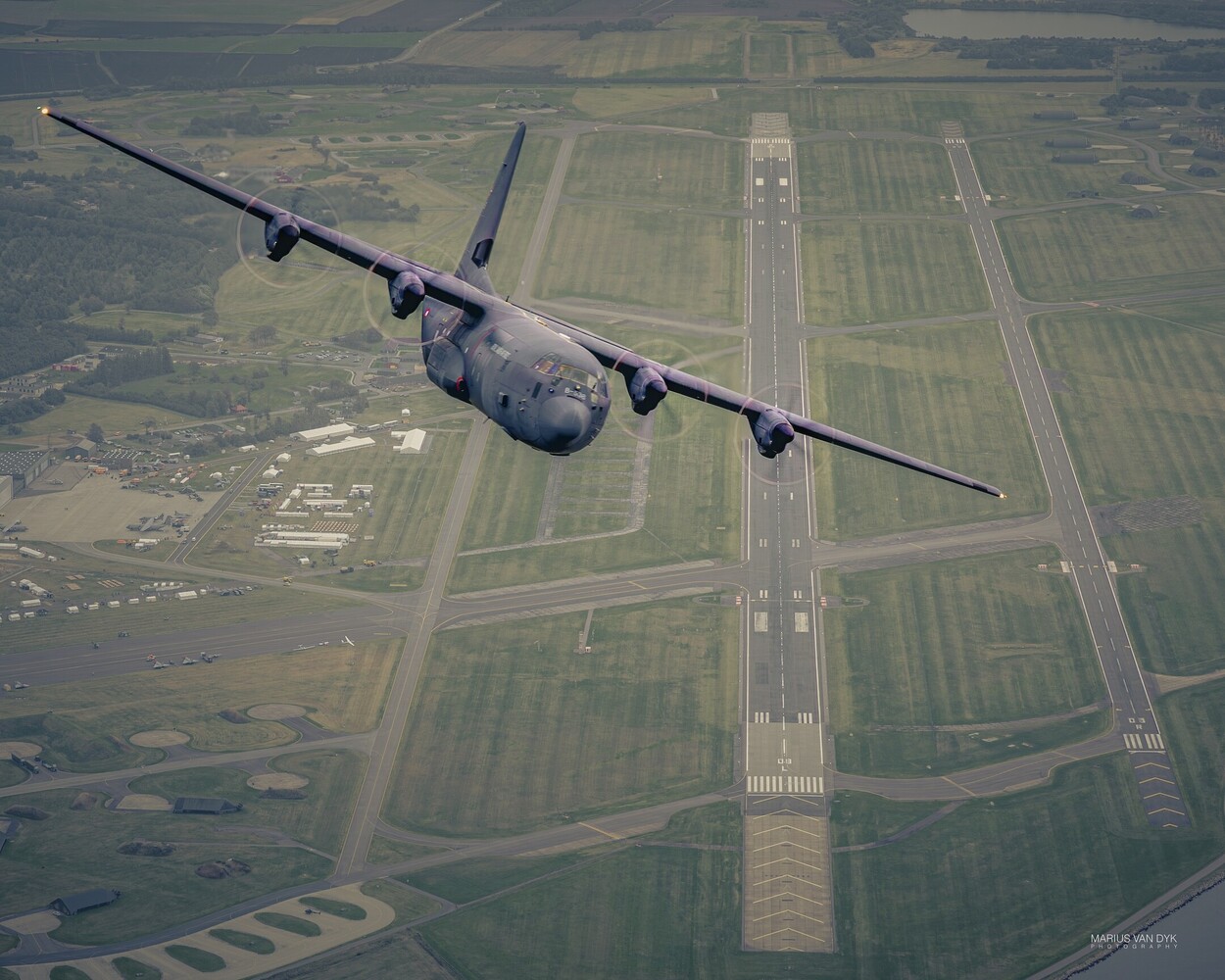Gray C-130 Hercules military transport plane flying low over a green airfield, its long runway stretching away below.