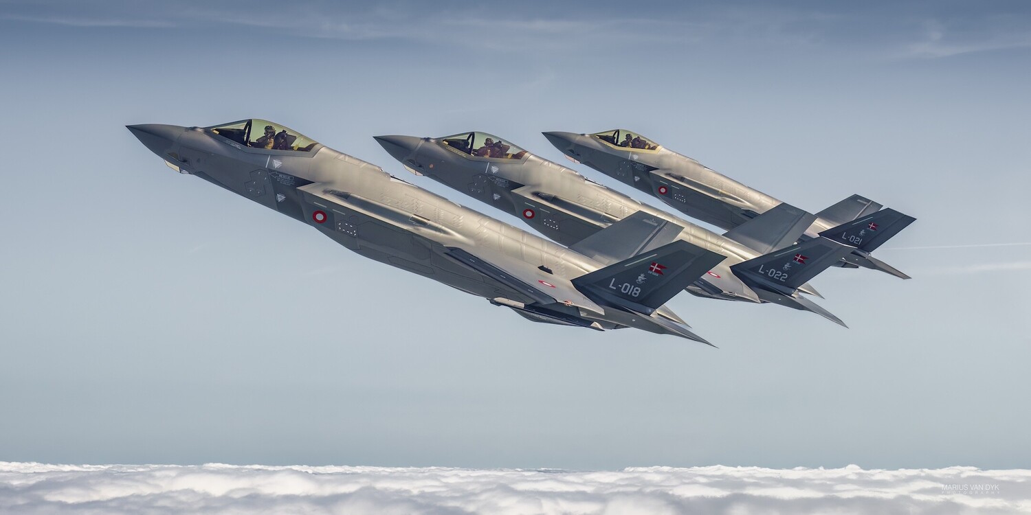 Three gray Danish F-35 Lightning II fighter jets flying in staggered formation above a cloud layer under a pale blue sky.