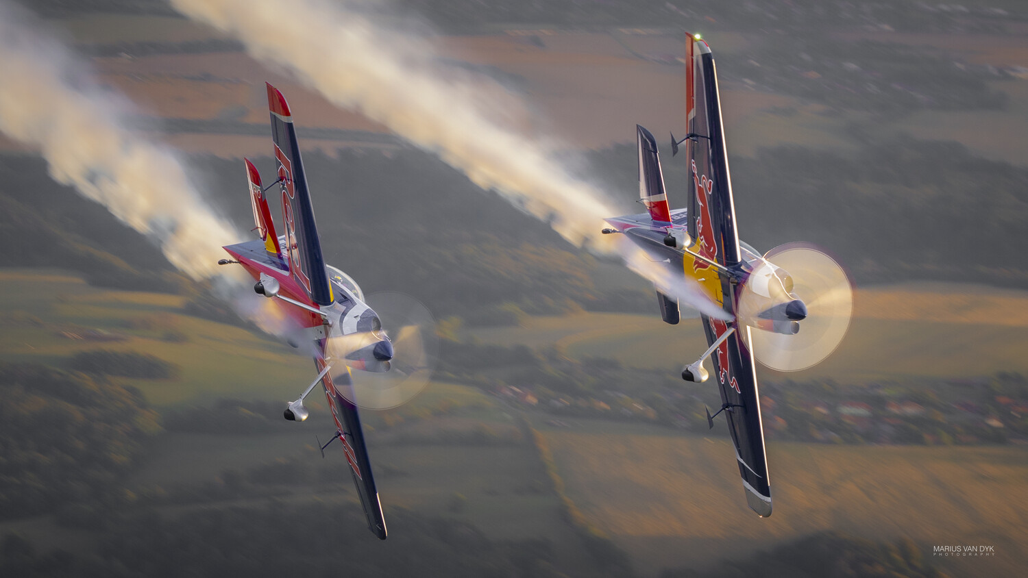 Two Red Bull-livery aerobatic aircraft flying in formation, trailing white smoke over farmland at sunset.