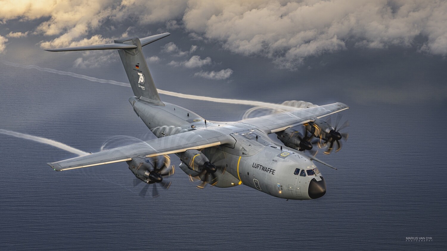 Grey Luftwaffe Airbus A400M military transport plane flying low over the sea, four propellers blurred, with contrails trailing behind