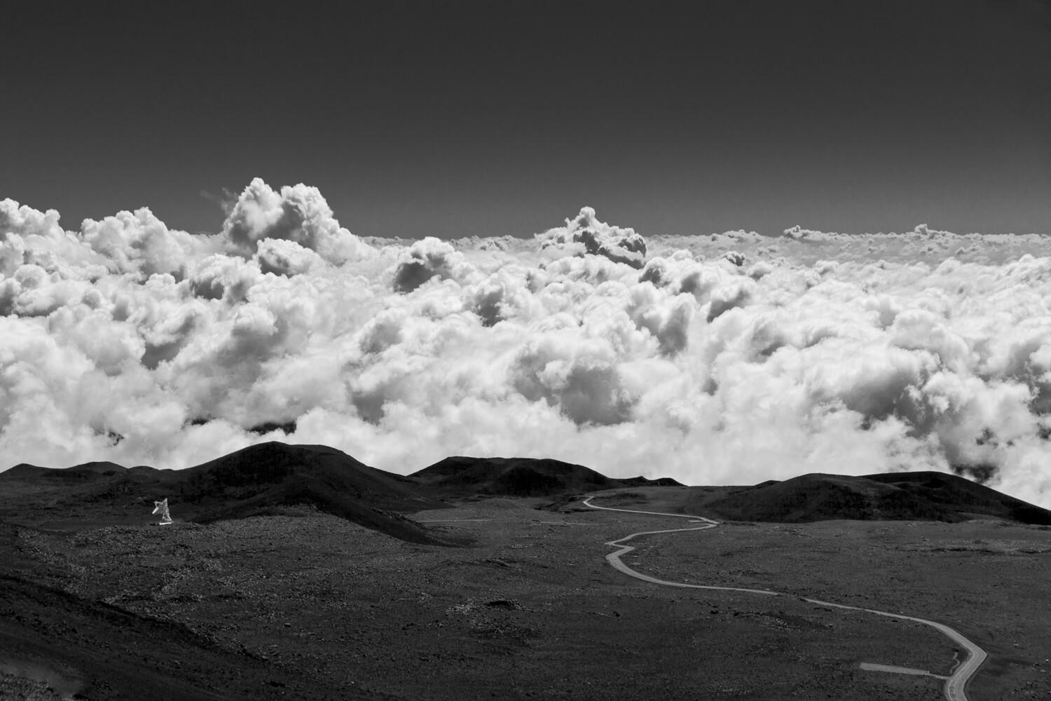 Black and white landscape showing dramatic cumulus clouds above a dark volcanic terrain with winding road