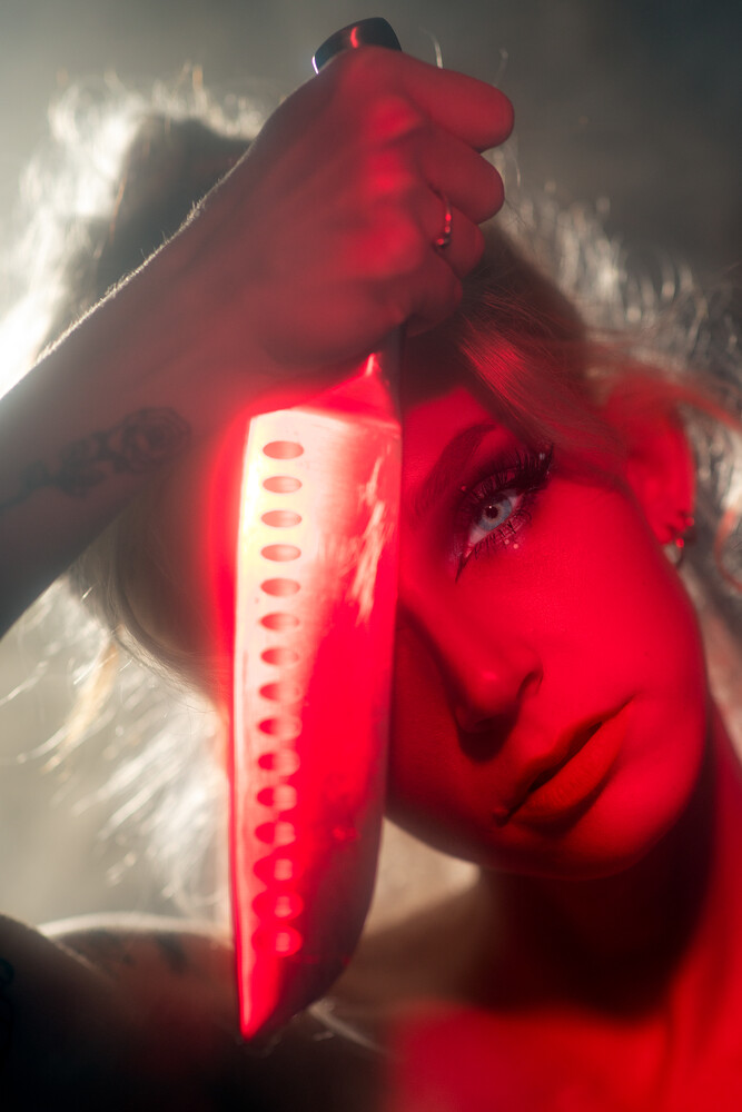 Woman's face washed in red light, eyes lowered, with a red-lit knife blade held upright beside her face against a hazy, smoky backdrop.