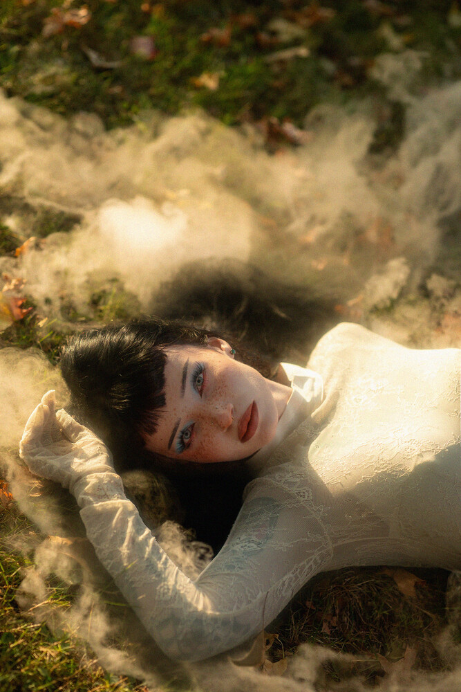 Woman with dark bobbed hair and red lips lies on mossy ground in a white lace long-sleeved top, surrounded by soft wispy smoke or mist.