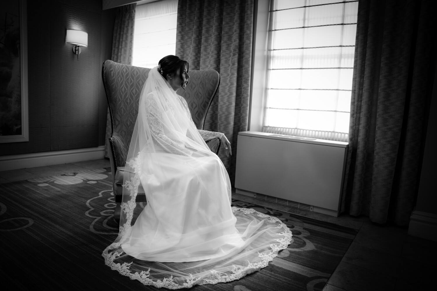 Black-and-white photo of a bride in a long veil seated in a wingback chair, gazing out a window in a softly lit room.