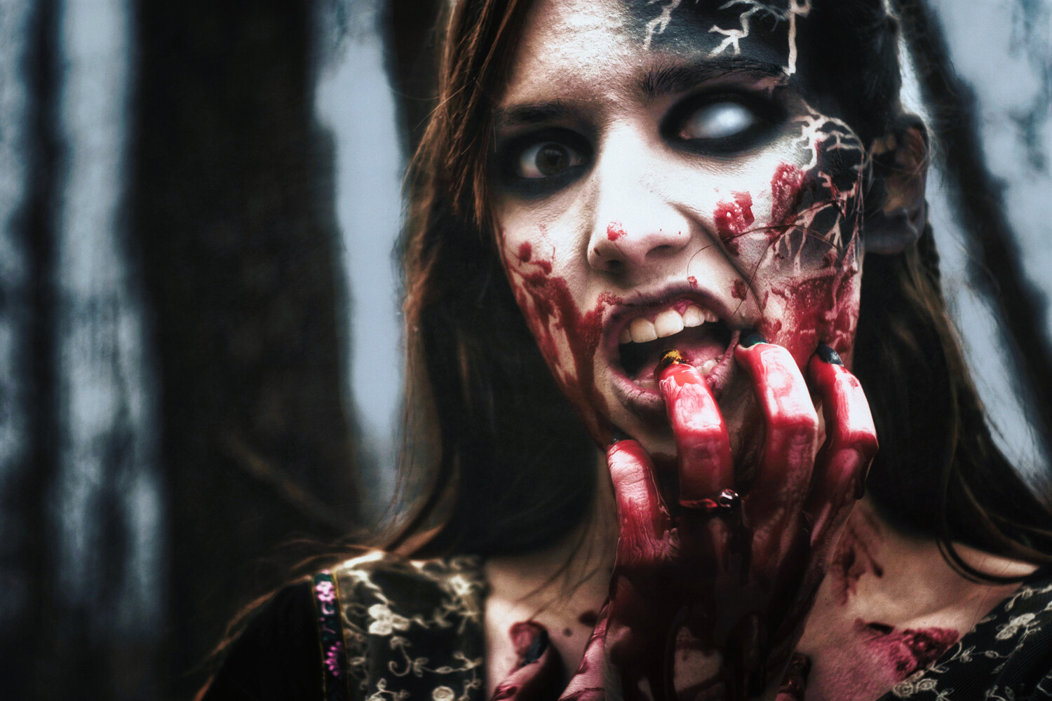 Woman in zombie makeup with blood-smeared face and hand, mouth open, staring wide-eyed in a blurred dark forest.