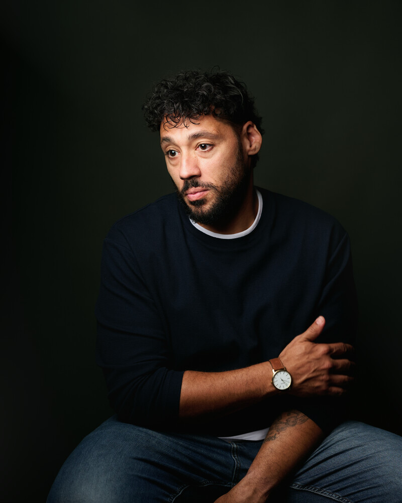 Man with curly hair and a short beard in a navy sweater and silver wristwatch, seated against a dark backdrop and gazing to the side.