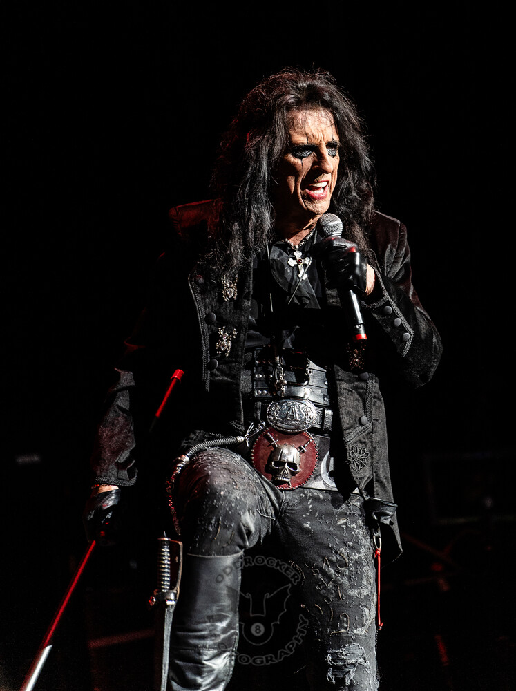 Male performer with long black hair singing into a handheld mic, holding a red-and-white cane against a dark stage