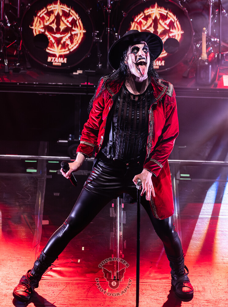 Performer in a black wide-brimmed hat and red jacket singing into a microphone under red stage lighting
