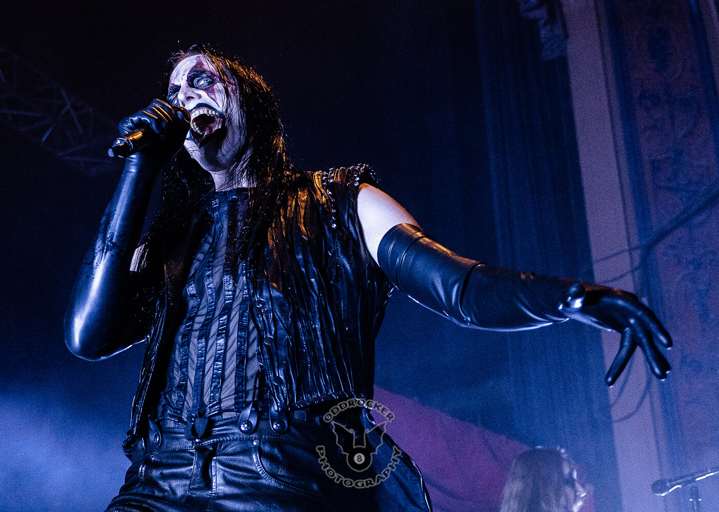 Vocalist in white corpse paint and black leather gloves singing into a microphone under blue stage lighting.