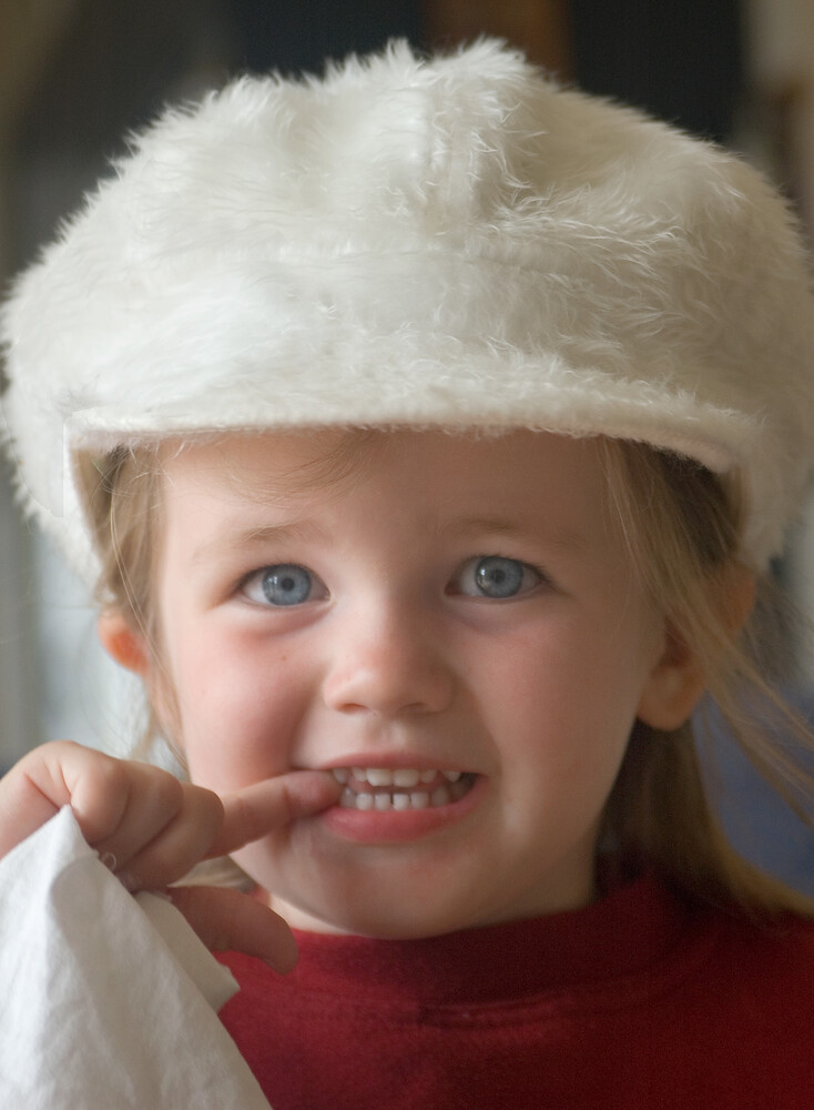 Young child wearing a white fuzzy winter hat and red sweater, smiling at camera with finger near mouth