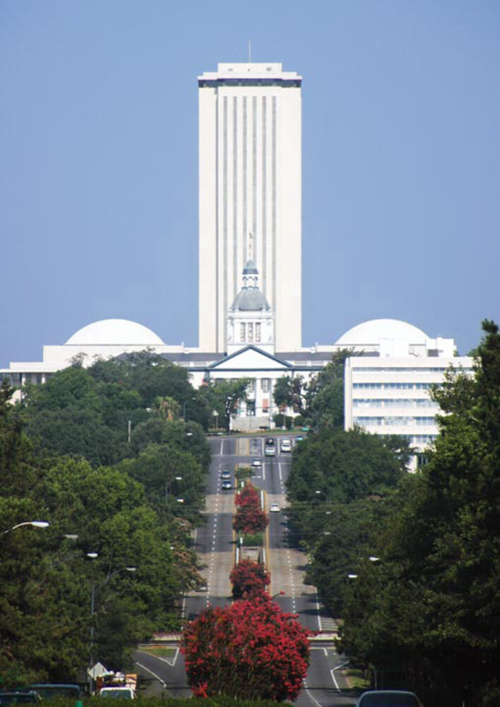 Tall modernist tower with vertical ribbing framed by symmetrical domed structures and tree-lined avenue