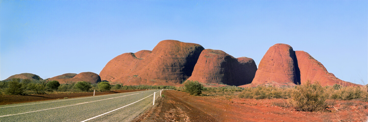 Red rock formations rise from the Australian outback along a straight desert highway