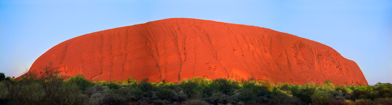 Uluru (Ayers Rock) glowing deep orange-red against clear blue sky with sparse desert vegetation below